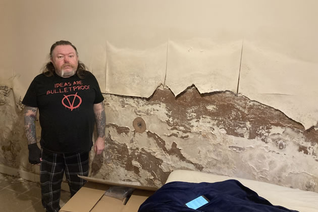 Michael Brady standing next to his bedroom wall where wallpaper has rotted and peeled off 